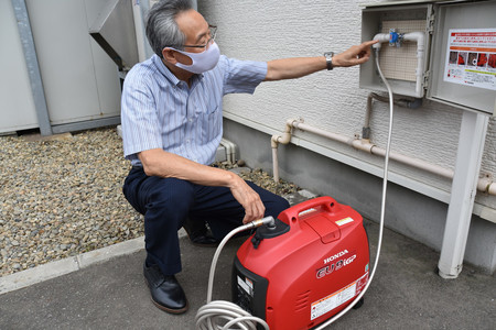 LPガス発電機とガス配管をつなぎ、始動手順を説明する障害者就労継続支援施設「One memory」の沢田修明さん=8月18日、秋田市